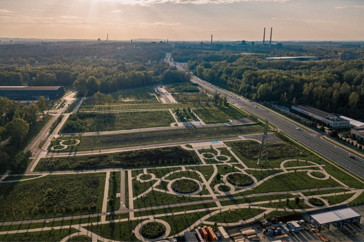 Widok z drona na tereny inwestycyjne z siecią dróg i zielenią, przy głównej arterii komunikacyjnej, w tle las i zabudowa.
