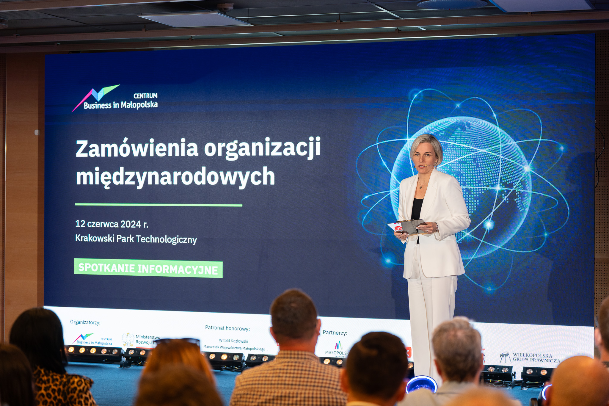 Granatowe tło z białym napisem Zamówienia organizacji międzynarodowych, kobieta ubrana w biały garnitur prowadzi spotkanie