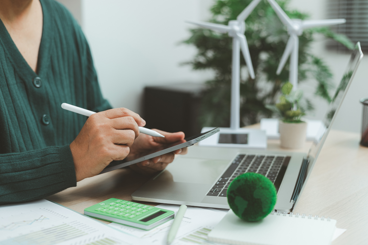 osoba pisząca na tablecie, w tle laptop na biurku, obok niego małe figurki wiatraków, przed laptopem kula imitująca kulę ziemską
